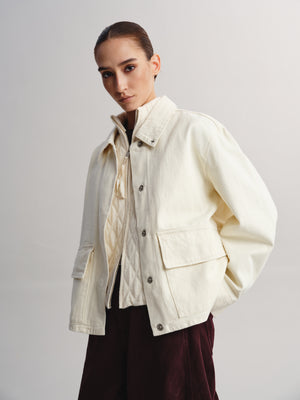 A model wearing a cream-colored utility jacket over a cream shirt and dark burgundy pants, photographed against a neutral light gray studio background.