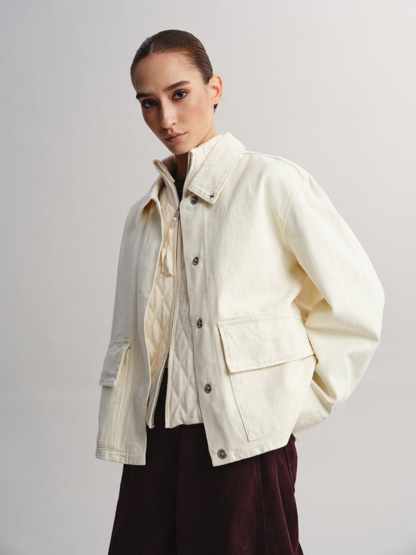 A model wearing a cream-colored utility jacket over a cream shirt and dark burgundy pants, photographed against a neutral light gray studio background.