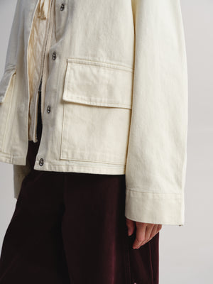 Close-up of a person wearing an off-white utility jacket with flap pockets over a cream shirt and dark burgundy trousers, against a plain light gray background.