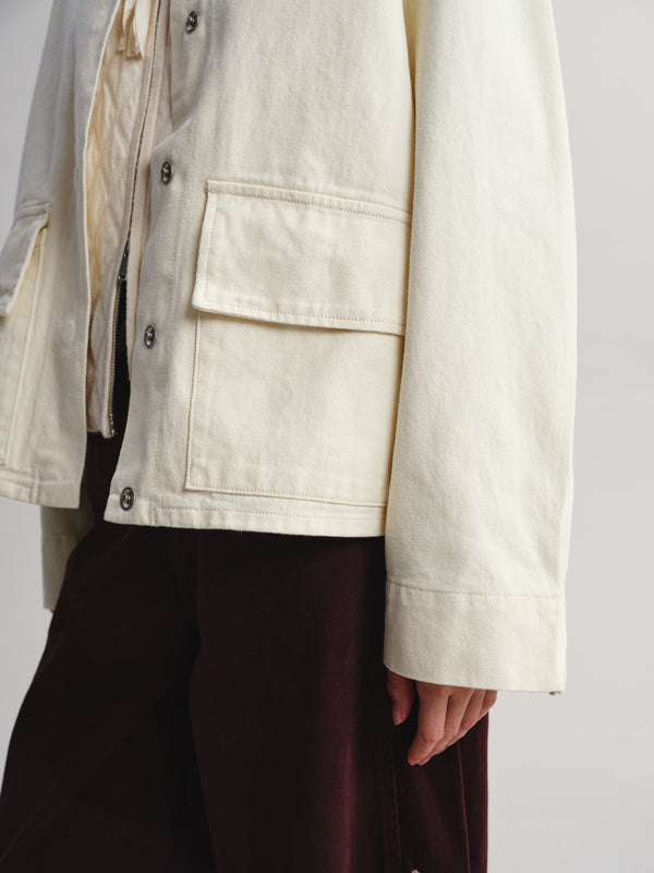 Close-up of a person wearing an off-white utility jacket with flap pockets over a cream shirt and dark burgundy trousers, against a plain light gray background.