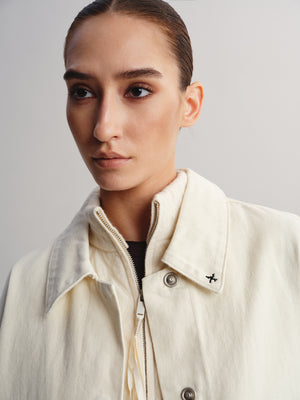 A close-up portrait of a model wearing a cream-colored textured jacket with a collar, zipper, and metal snap buttons, set against a plain light gray background.