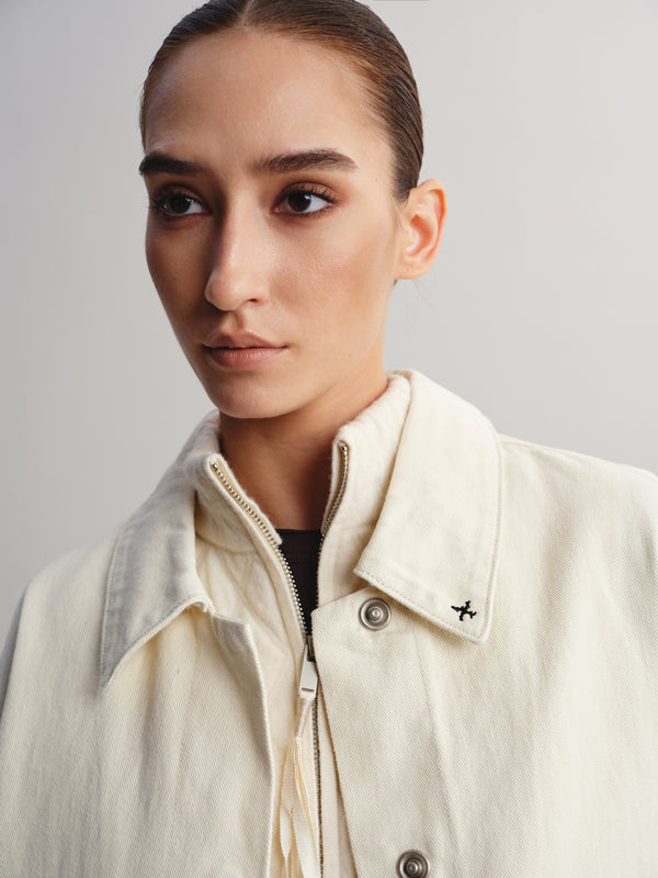 A close-up portrait of a model wearing a cream-colored textured jacket with a collar, zipper, and metal snap buttons, set against a plain light gray background.