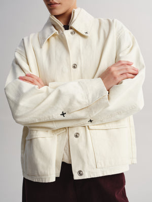 A person wearing a cream-colored canvas utility jacket with silver buttons and small black embroidered cross details on the collar and cuffs, paired with dark burgundy pants against a light gray background.