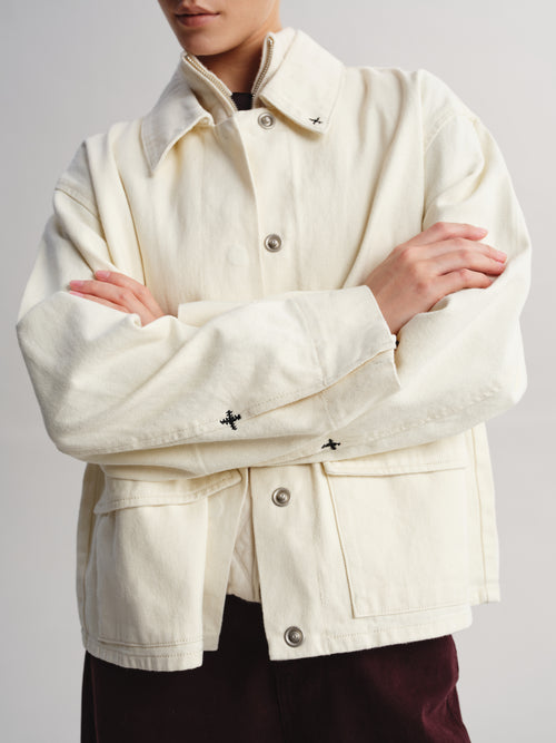 A person wearing a cream-colored canvas utility jacket with silver buttons and small black embroidered cross details on the collar and cuffs, paired with dark burgundy pants against a light gray background.