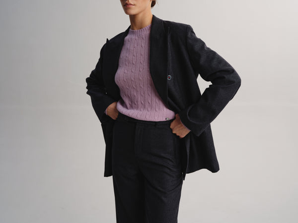 A model wearing a dark charcoal gray double-breasted blazer over a lavender cable-knit sweater and dark trousers, standing against a light gray studio background.