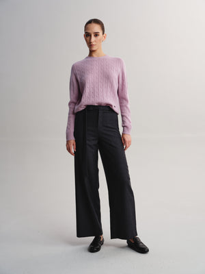 A model stands against a light gray studio background wearing a dusty pink cable-knit sweater, black wide-leg trousers, and black loafers.
