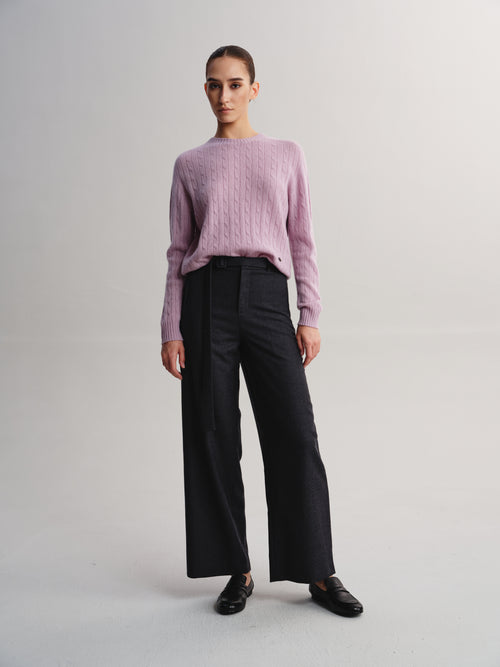 A model stands against a light gray studio background wearing a dusty pink cable-knit sweater, black wide-leg trousers, and black loafers.