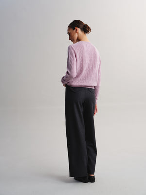 A woman is shown from the back wearing a light pink cable-knit sweater and dark charcoal wide-leg trousers, standing against a neutral light gray studio background.