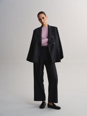 A model stands against a light gray studio background wearing a black double-breasted blazer, matching wide-leg trousers, a light pink ribbed top, and black loafers.