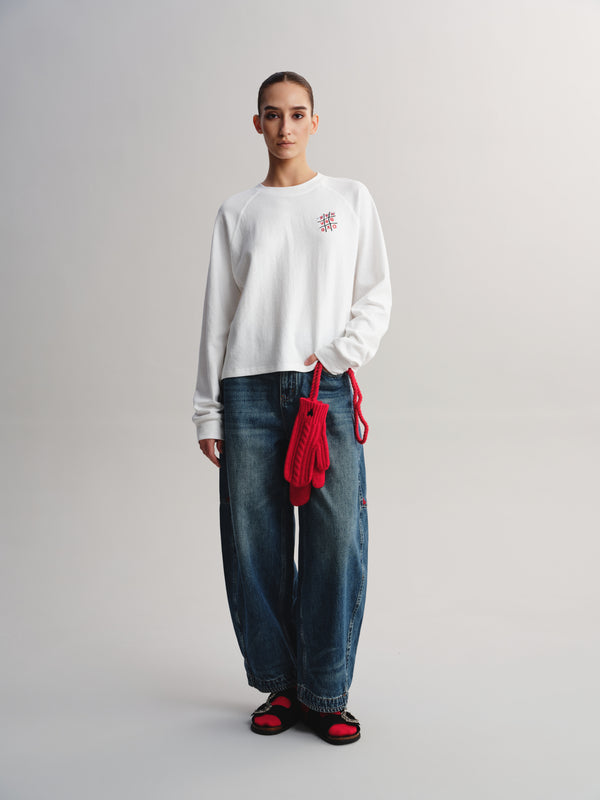 A model stands against a light gray studio background wearing a white crewneck sweatshirt with a small pink embroidered logo on the left chest, loose blue jeans, and red-and-black sandals. A bright red knitted bag is draped over the hip.