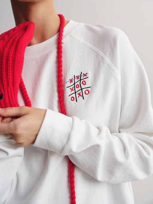 A person wearing a white crewneck sweatshirt with a small red and black tic-tac-toe graphic on the left chest, styled with a bright red knit scarf against a light gray background.
