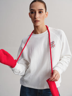 A woman wearing a white crewneck sweatshirt with a red embroidered tic-tac-toe graphic on the left chest, styled with a red braided lanyard and red knit accessories against a light gray studio background.