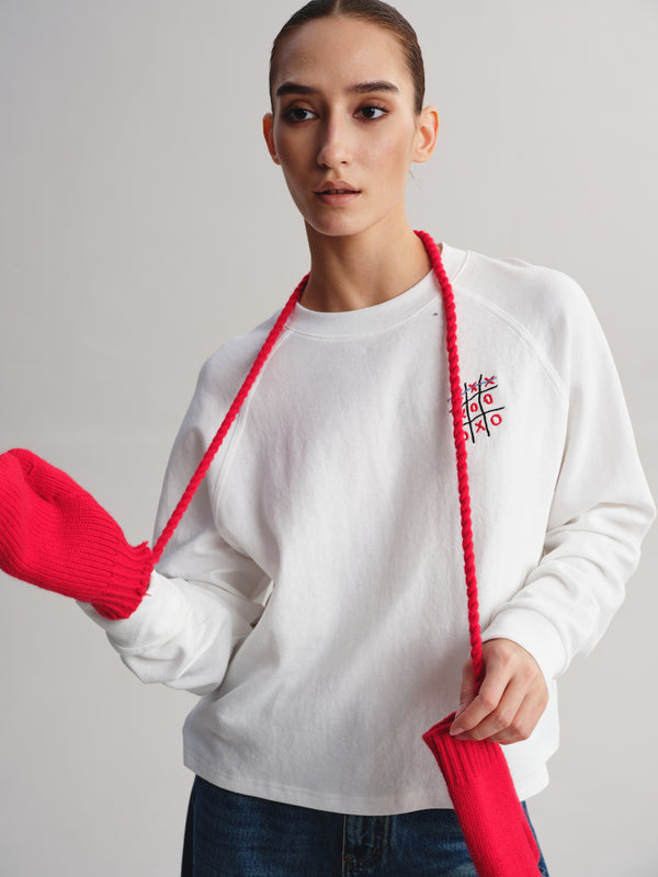 A woman wearing a white crewneck sweatshirt with a red embroidered tic-tac-toe graphic on the left chest, styled with a red braided lanyard and red knit accessories against a light gray studio background.