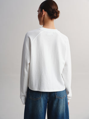 A woman shown from behind wearing a white long-sleeve raglan sweatshirt and blue jeans against a plain light gray studio background.