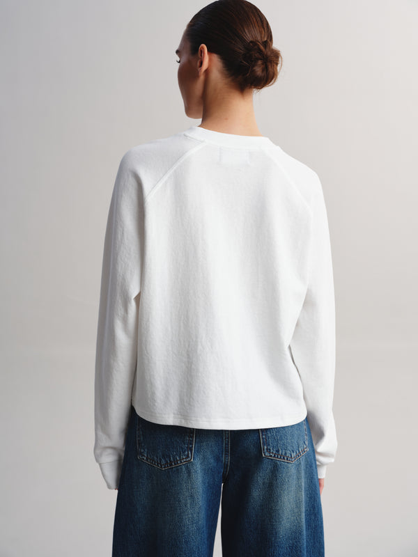 A woman shown from behind wearing a white long-sleeve raglan sweatshirt and blue jeans against a plain light gray studio background.