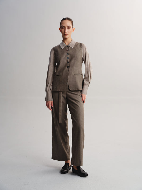 A model stands against a light gray studio background wearing a coordinated taupe-brown two-piece suit with a collared jacket and wide-leg trousers, paired with black loafers.