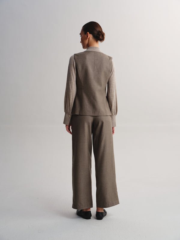 A model viewed from the back wearing a matching taupe-brown two-piece suit consisting of a long-sleeve blazer and wide-leg trousers, paired with black shoes against a light gray studio background.