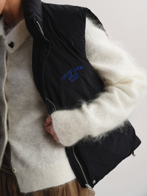 A close-up of a person wearing a black quilted puffer vest over a fuzzy cream knit sweater. The vest has blue embroidered text reading 'CHLOE CHEN CLUB' on the chest and silver zippers.