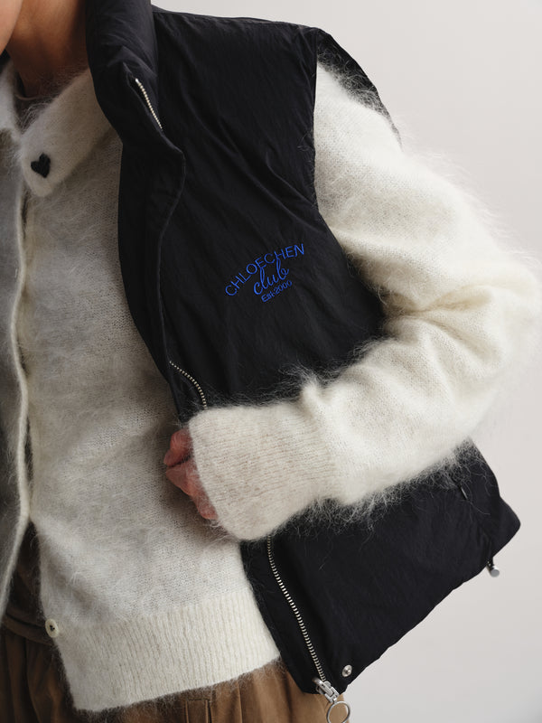 A close-up of a person wearing a black quilted puffer vest over a fuzzy cream knit sweater. The vest has blue embroidered text reading 'CHLOE CHEN CLUB' on the chest and silver zippers.