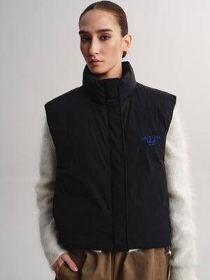 A model wearing a black sleeveless puffer vest with a high collar and blue embroidered text on the chest, layered over a fuzzy cream sweater and brown pants, against a light gray studio background.