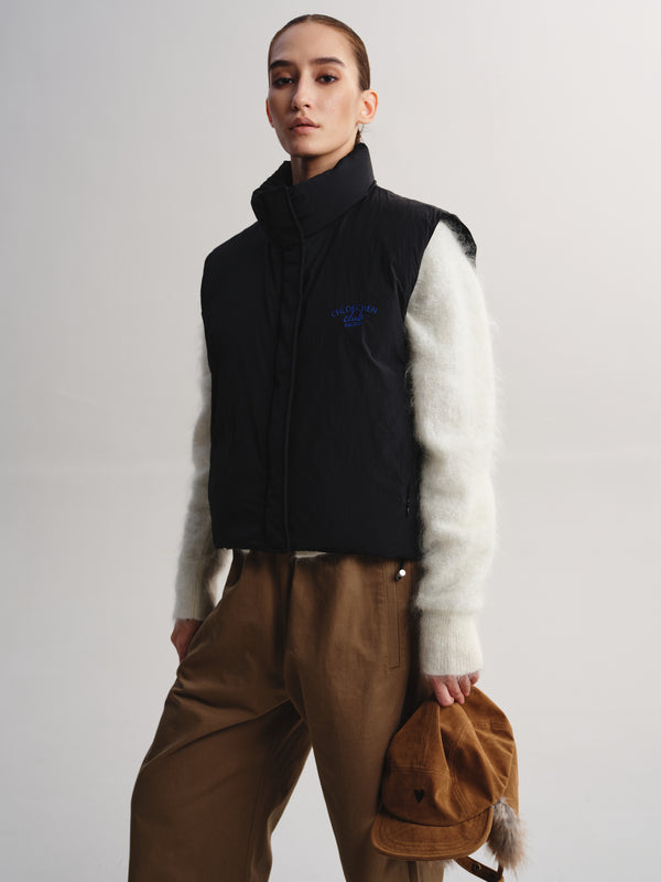 A model wearing a black sleeveless puffer vest with a high collar, featuring a blue embroidered logo on the chest, layered over a fuzzy white long-sleeve top and paired with brown pants, holding a brown corduroy cap against a light gray studio background.