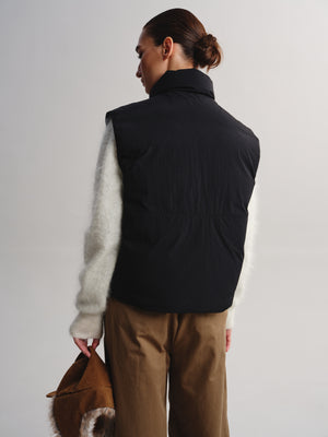 A model shown from the back wearing a black sleeveless puffer vest over a fuzzy white long-sleeve top and brown pants, holding a brown fur-trimmed bag against a light gray studio background.