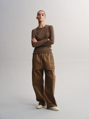 A model stands against a light gray studio background wearing a brown long-sleeve crewneck top and matching brown wide-leg pants, paired with white slip-on shoes.