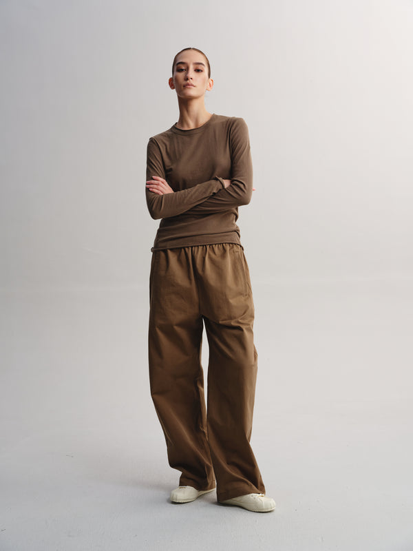 A model stands against a light gray studio background wearing a brown long-sleeve crewneck top and matching brown wide-leg pants, paired with white slip-on shoes.