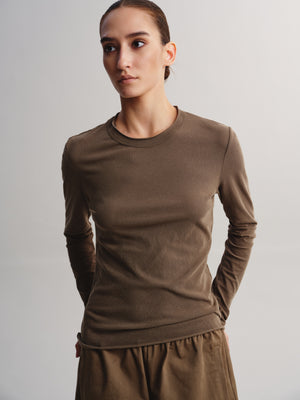 A woman modeling a fitted, long-sleeve crewneck top in a solid taupe-brown color, paired with matching brown bottoms, against a neutral light gray studio background.