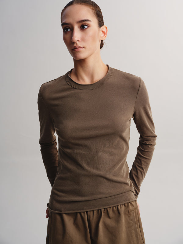 A woman modeling a fitted, long-sleeve crewneck top in a solid taupe-brown color, paired with matching brown bottoms, against a neutral light gray studio background.
