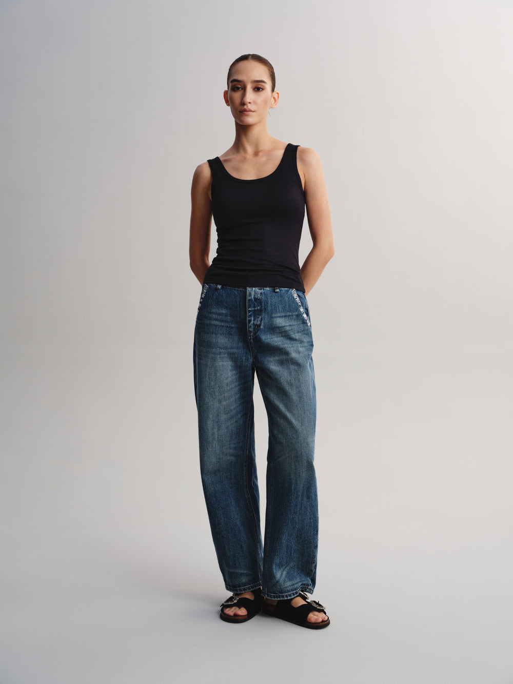 A close-up of a model wearing a black ribbed scoop-neck tank top paired with blue jeans against a light gray background.