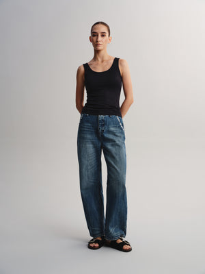 A woman stands against a light gray studio background wearing a black sleeveless tank top, blue wide-leg jeans, and black sandals.