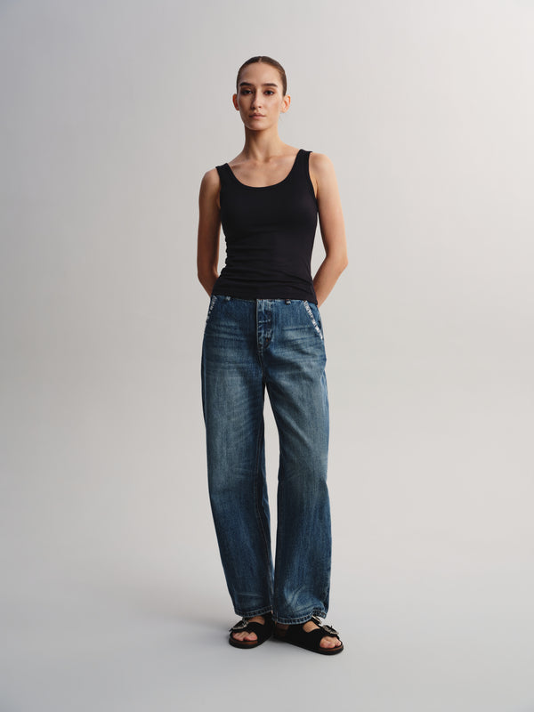 A woman stands against a light gray studio background wearing a black sleeveless tank top, blue wide-leg jeans, and black sandals.