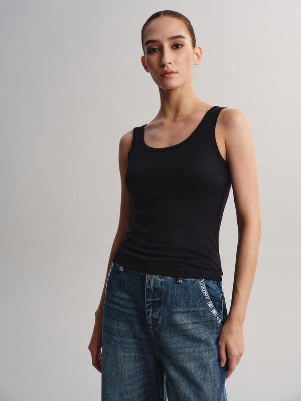 A model wearing a black scoop-neck tank top and blue jeans, standing against a neutral light gray studio background.