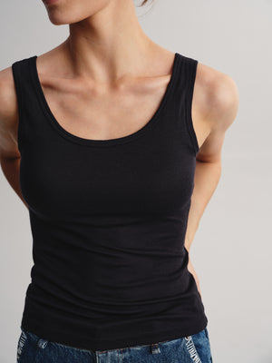 A close-up of a model wearing a black ribbed scoop-neck tank top paired with blue jeans against a light gray background.