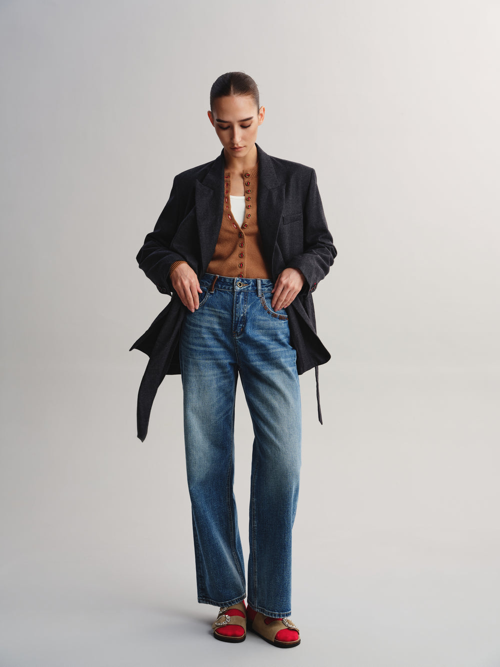 A model stands against a light gray studio background wearing a black blazer over a brown textured top, blue wide-leg jeans, and red-and-brown flat shoes.