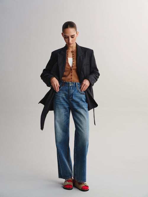 A model stands against a light gray studio background wearing a black blazer over a brown textured top, blue wide-leg jeans, and red-and-brown flat shoes.