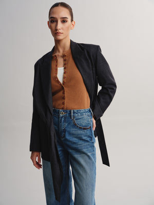 A model wearing a black textured blazer over a brown button-up cardigan, a white inner top, and blue jeans, photographed against a neutral light gray studio background.