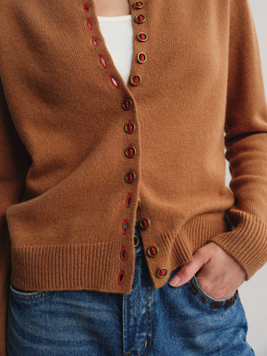 A close-up of a person wearing a camel brown knit cardigan with red and gold decorative buttons, paired with a white top and blue jeans.