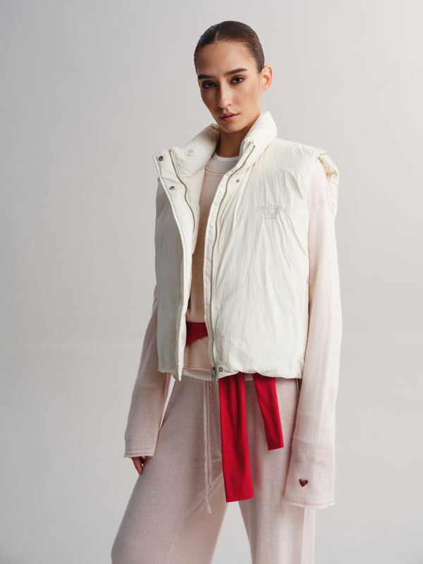 A model wearing a cream-colored sleeveless puffer vest over a light pink long-sleeve top and matching pink sweatpants, with a bright red tie belt. The photo is taken against a neutral light gray studio background.