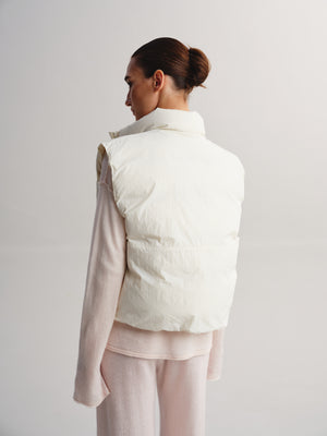 A model shown from the back wearing a cream-colored puffer vest over a light pink long-sleeve top and matching pink pants, set against a plain light gray studio background.
