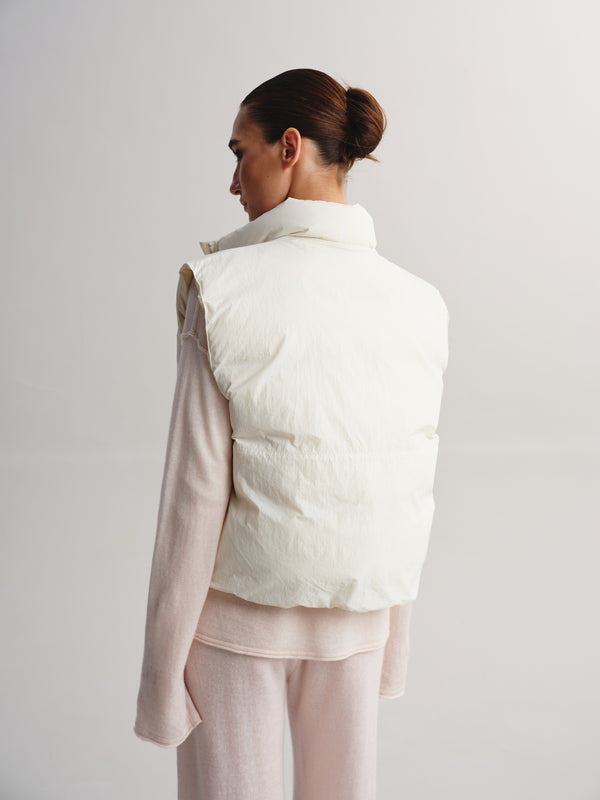A model shown from the back wearing a cream-colored puffer vest over a light pink long-sleeve top and matching pink pants, set against a plain light gray studio background.