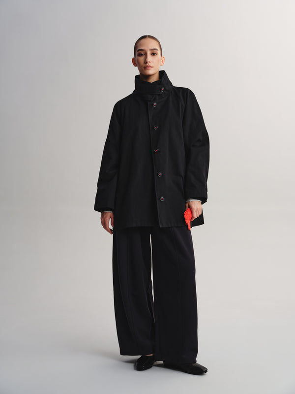 A model stands against a light gray studio background wearing a black high-neck button-front coat with wide-leg black pants and black shoes, holding a small bright red object in one hand.