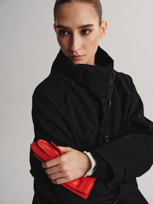 A model wearing a black high-collar jacket and holding a bright red clutch bag against a light gray studio background.