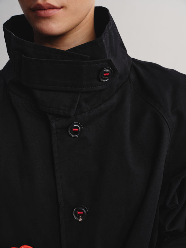 Close-up of a person wearing a black high-collar jacket with dark buttons that have a small red logo detail, against a light gray background.