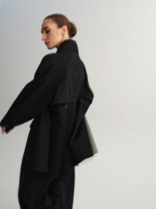 A model wearing an oversized black coat and matching wide-leg trousers, photographed against a plain light gray studio background.