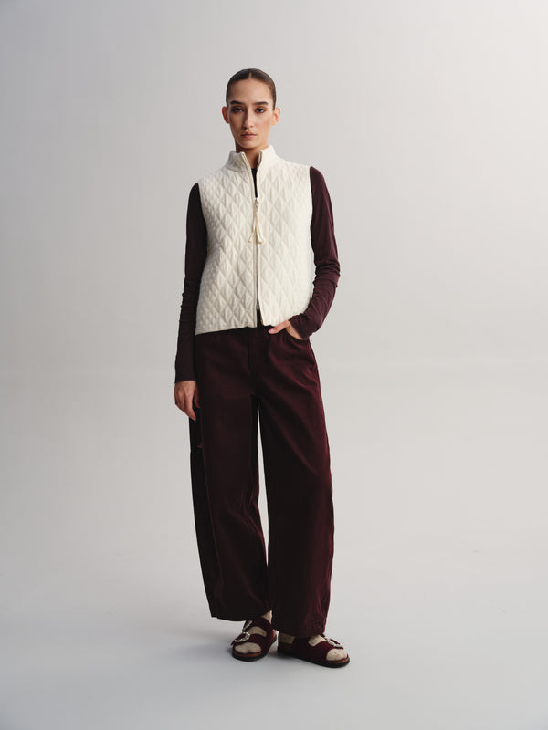 A model stands against a light gray studio background wearing a cream quilted zip-front vest over a dark burgundy long-sleeve top and matching wide-leg trousers, with dark sandals.