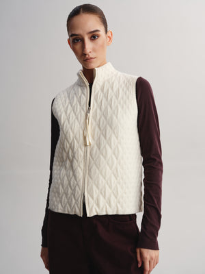 A model wearing a cream-colored quilted zip-up vest over a dark burgundy long-sleeve top and matching dark trousers, photographed against a neutral light gray studio background.