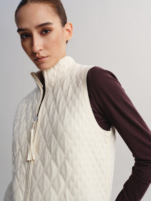 A close-up of a model wearing a cream-colored quilted zip-up vest over a dark burgundy long-sleeve shirt, set against a plain light gray studio background.