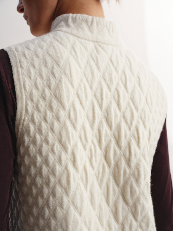 Close-up back view of a person wearing a quilted cream-colored sleeveless vest over a dark plum long-sleeve top.